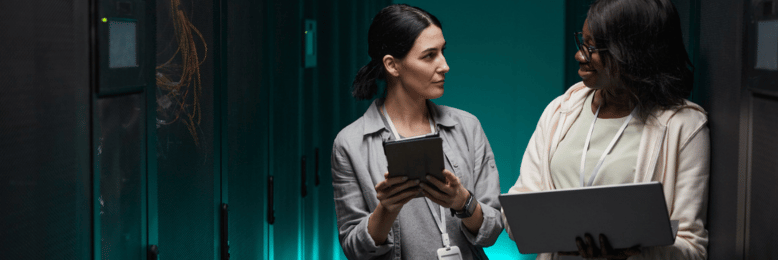 Female IT engineers with a laptop and tablet discussing data backup in a server room