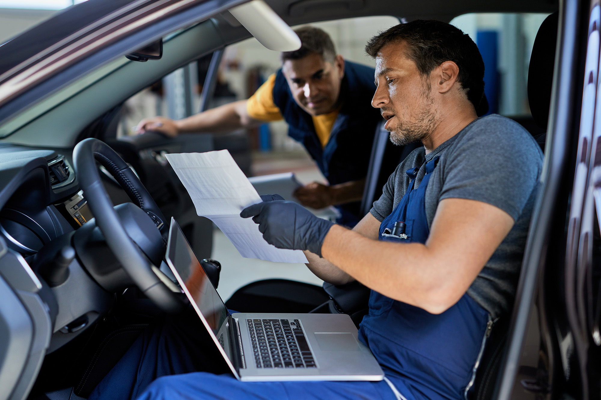 auto_how_step2 (Auto repairman talking to his colleague while running car diagnostic and analyzing data in a workshop)