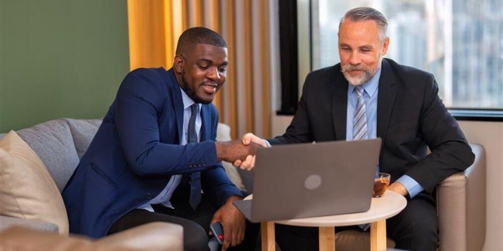 Two professionals shaking hands while looking at a laptop, discussing how vCIOs and MSPs can work together