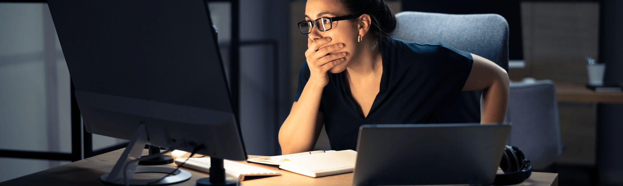 A woman at her desk looks shocked, covering her mouth as she stares at her computer, possibly reacting to an unexpected issue or data loss.