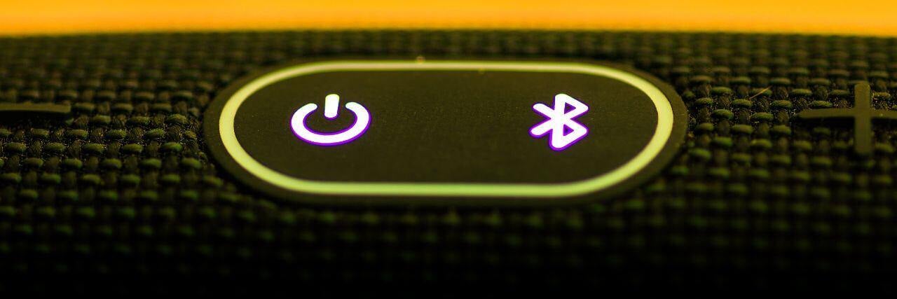 A close-up of a Bluetooth and power button on a black textured device, illuminated with purple light, representing wireless connectivity and device control.