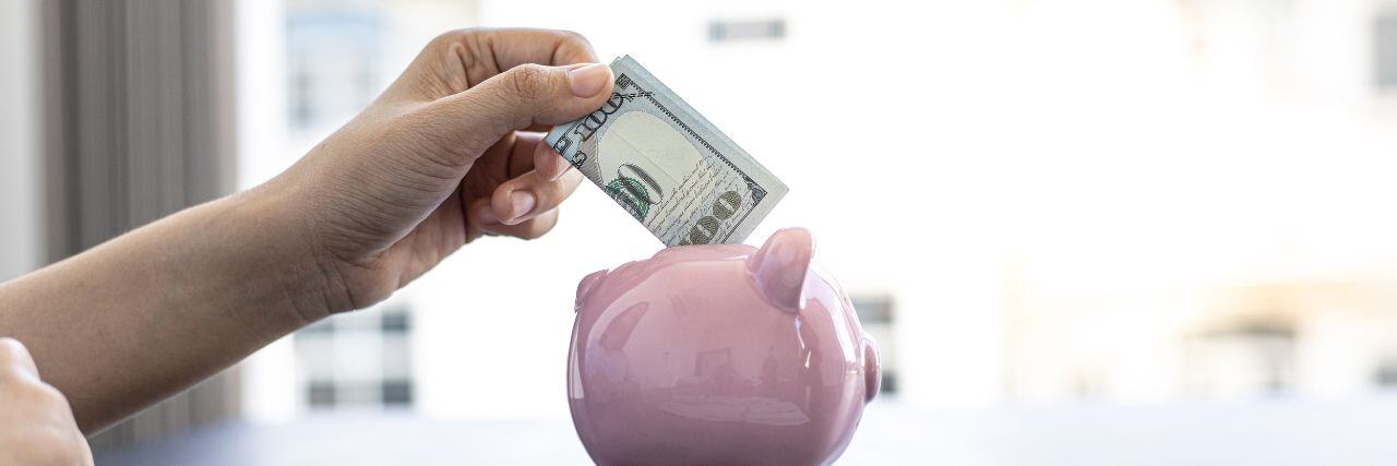 a hundred dollar bill folded being inserted in a pink piggy bank